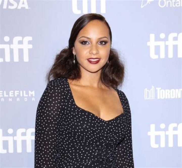 Jasmine Cephas Jones smiling in a dark blue V-neck dress and silver earrings.